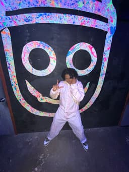 kidd stands, gap-legged, in hooded white coveralls in front of a smiley-face character. she makes an "L" with her left hand