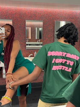 nunu takes a mirror selfie while kidd has her back to the mirror. the back of kidd's WiseOne shirt reads, "SORRY BABY I GOT TO HUSTLE