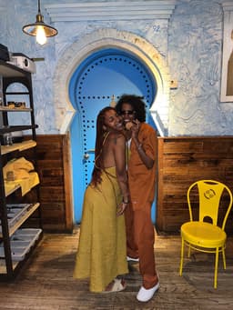 kidd poses with girlfriend, nunu, in front of a blue Moorish door at a downtown NYC restaurant. they both smile. kidd, wearing shades, holds her mother's gold medallion near her face